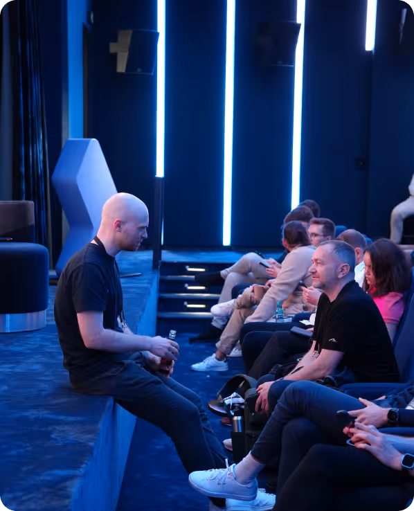 A young man in black sits on a stage edge holding a bottle, talking to a seated group of people in a blue-lit room.