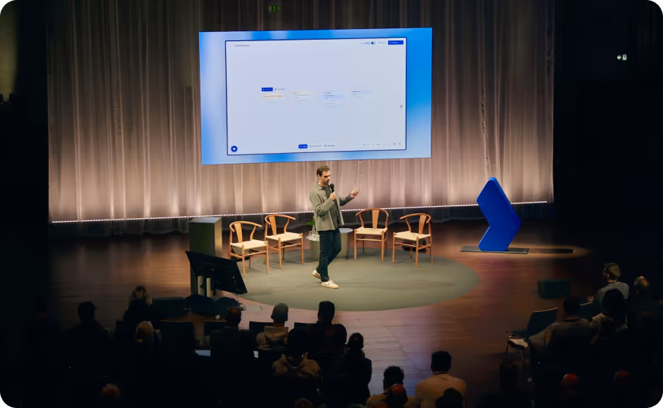Man holding a microphone presenting on stage with chairs behind him and a large screen displaying a software interface.