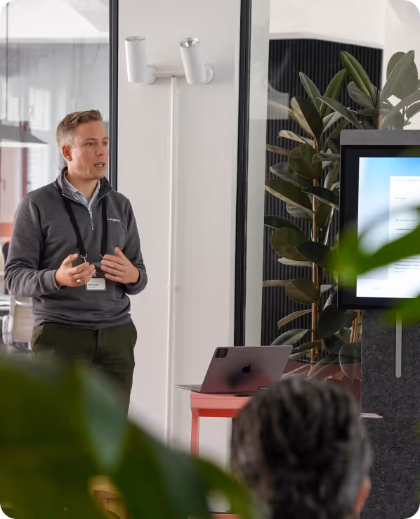 A man wearing a gray pullover and name badge speaking in front of a seated audience with a laptop and large screen in a modern office setting.